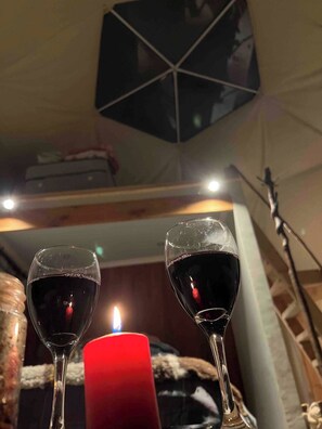Zwei Weingläser auf einem Tisch mit einer brennenden Kerze, einer geometrischen Deckenleuchte und einem Blick auf ein Bett mit Decke.