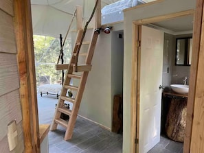A wooden ladder leaning against a wall in a room with a tiled floor, a door leading to a bathroom with a sink, and a view of the outdoors.