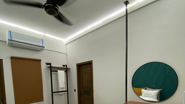 A bedroom with a bed, a ceiling fan, an air conditioning unit, a towel rack, a wooden door, a mirror with a circular frame, and a bedside table. - Lovely 1-bedroom apartment with AC in vibrant Karachi (Karachi)