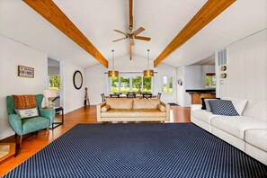 Un salón espacioso con techo alto, vigas de madera, un sofá grande, una mesa de centro y un comedor con vistas al exterior.