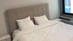 Una cama bien hecha con cabecera, una mesita de noche con cajón y una ventana que da a vistas de edificios.