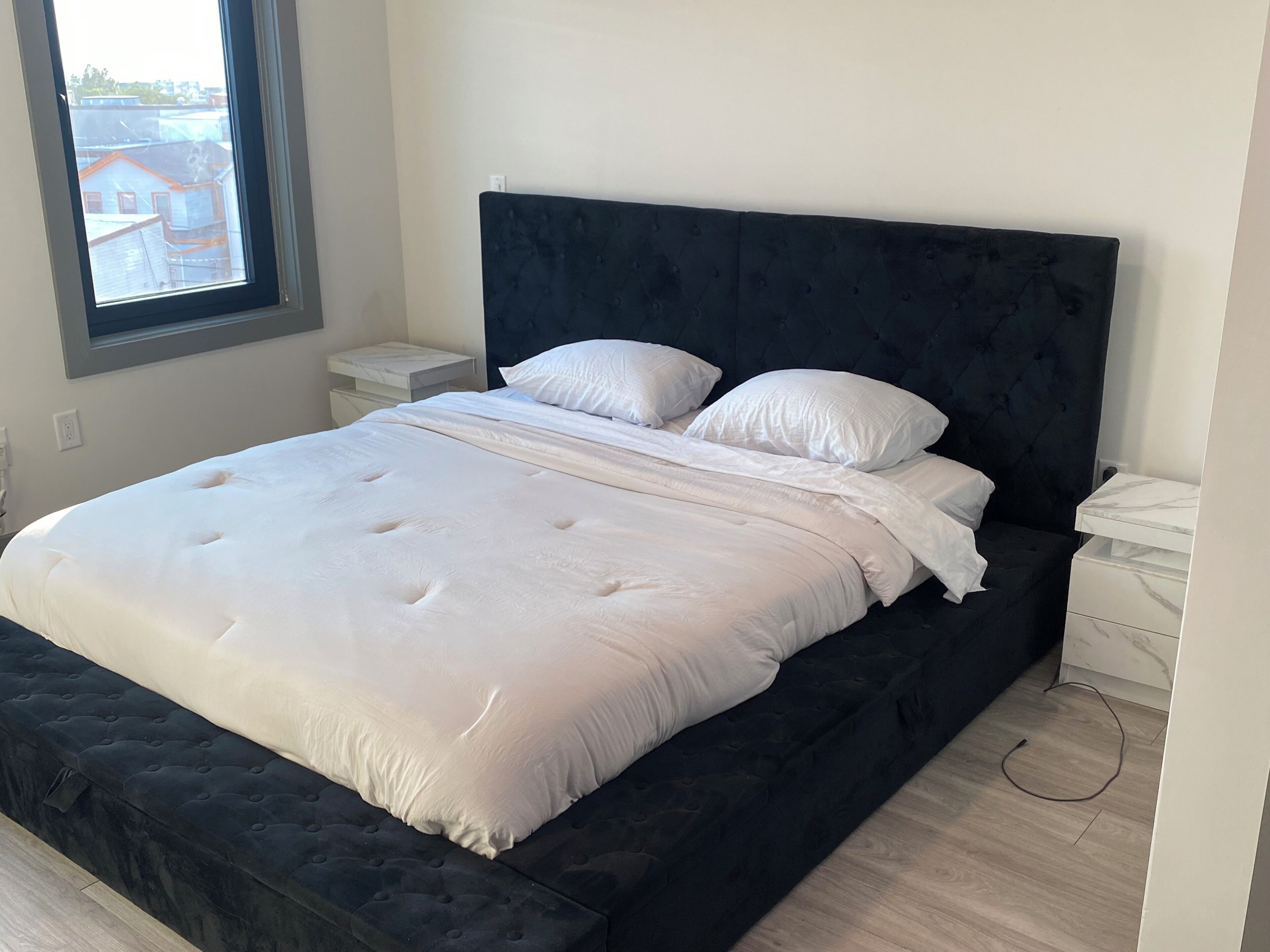 Un lit bien fait, avec une tête de lit sombre et du linge de lit blanc, une petite table de chevet et une fenêtre donnant sur des immeubles.