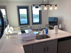 A modern kitchen with a marble countertop, a dishwasher, a sink, and a window with a view of a building and trees.