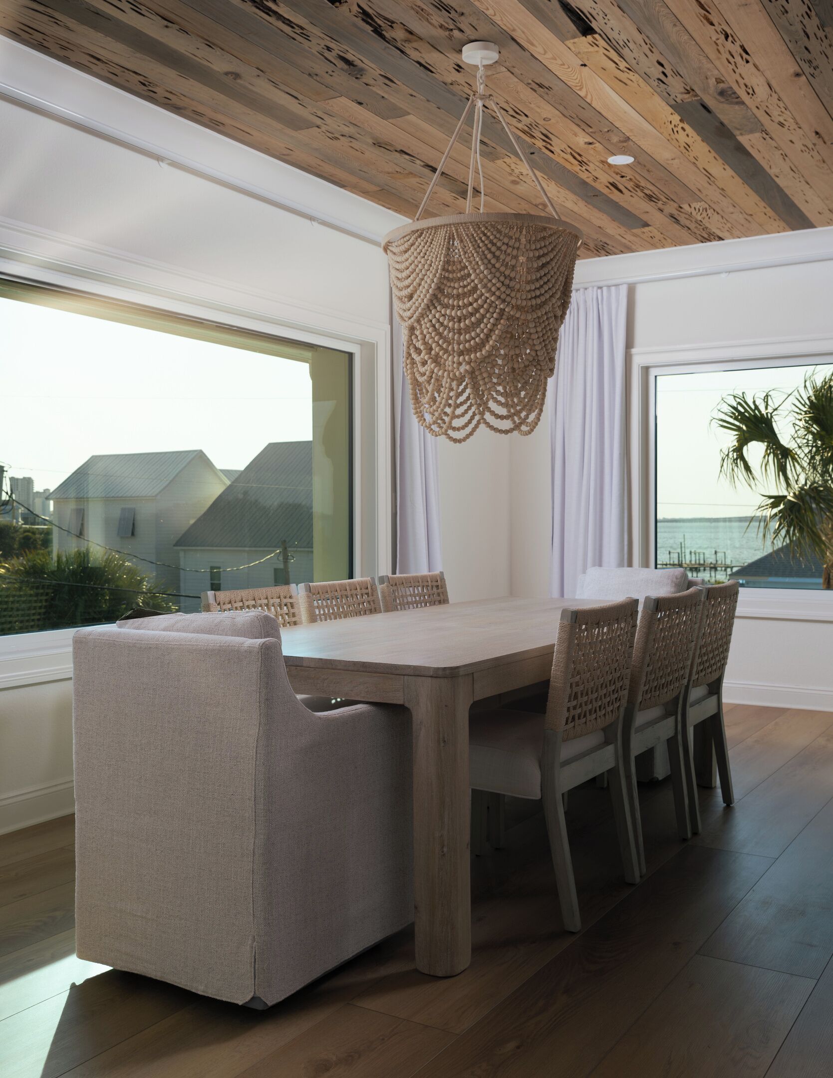 Ein zeitgemäßer Essbereich mit Holztisch und Stühlen, einer gewebten Pendelleuchte und großen Fenstern mit Meerblick.