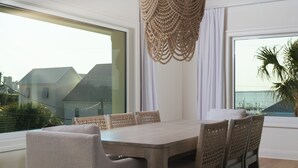 A modern dining area with a wooden table and chairs, a woven pendant light, and large windows offering a view of the sea. - New Stunning Luxury Beach Home with Private Pool! (Pensacola Beach)