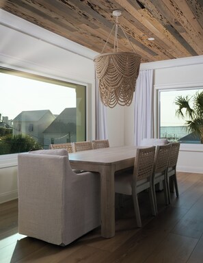 Ein zeitgemäßer Essbereich mit Holztisch und Stühlen, einer gewebten Pendelleuchte und großen Fenstern mit Meerblick.