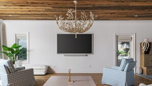 A living room with a wooden ceiling, a flat-screen TV, two checkered sofas, a coffee table, and a large plant. - New Stunning Luxury Beach Home with Private Pool! (Pensacola Beach)