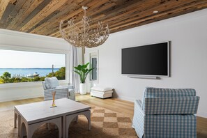 A modern living room with a wooden ceiling, a large window with a view, a chandelier, and a flat-screen TV. - New Stunning Luxury Beach Home with Private Pool! (Pensacola Beach)