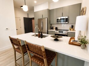 A modern kitchen with a central island, stainless steel appliances, and wooden chairs. - The Jardin Birch Suite | Near Downtown (Savannah)