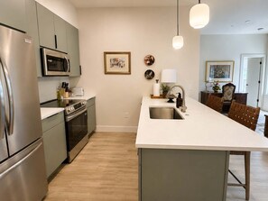 A modern kitchen with stainless steel appliances, a white countertop, and a wooden dining area. - The Jardin Birch Suite | Near Downtown (Savannah)