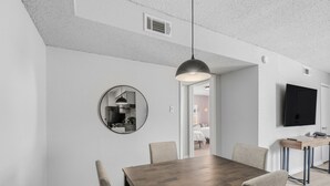 Uma sala de jantar com uma mesa de madeira, cadeiras, uma luminária pendente, um espelho redondo e uma TV na parede.