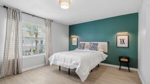 Quarto com cama, janela com cortinas, luminária fixada na parede e quadro na parede.