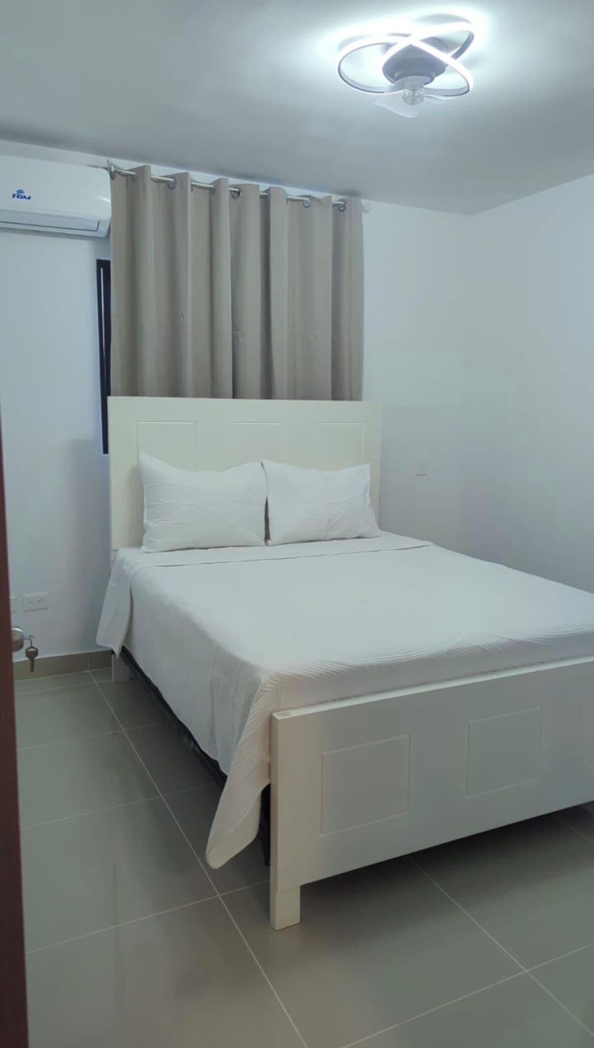 A bedroom with a bed, curtains, an air conditioning unit, and a ceiling light fixture.