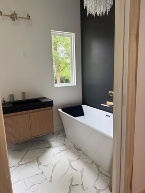 Ein Badezimmer mit einer weißen Badewanne, einem hölzernen Waschtisch, einem Marmorboden, einem Fenster mit Blick auf Bäume und einer Chandelier.