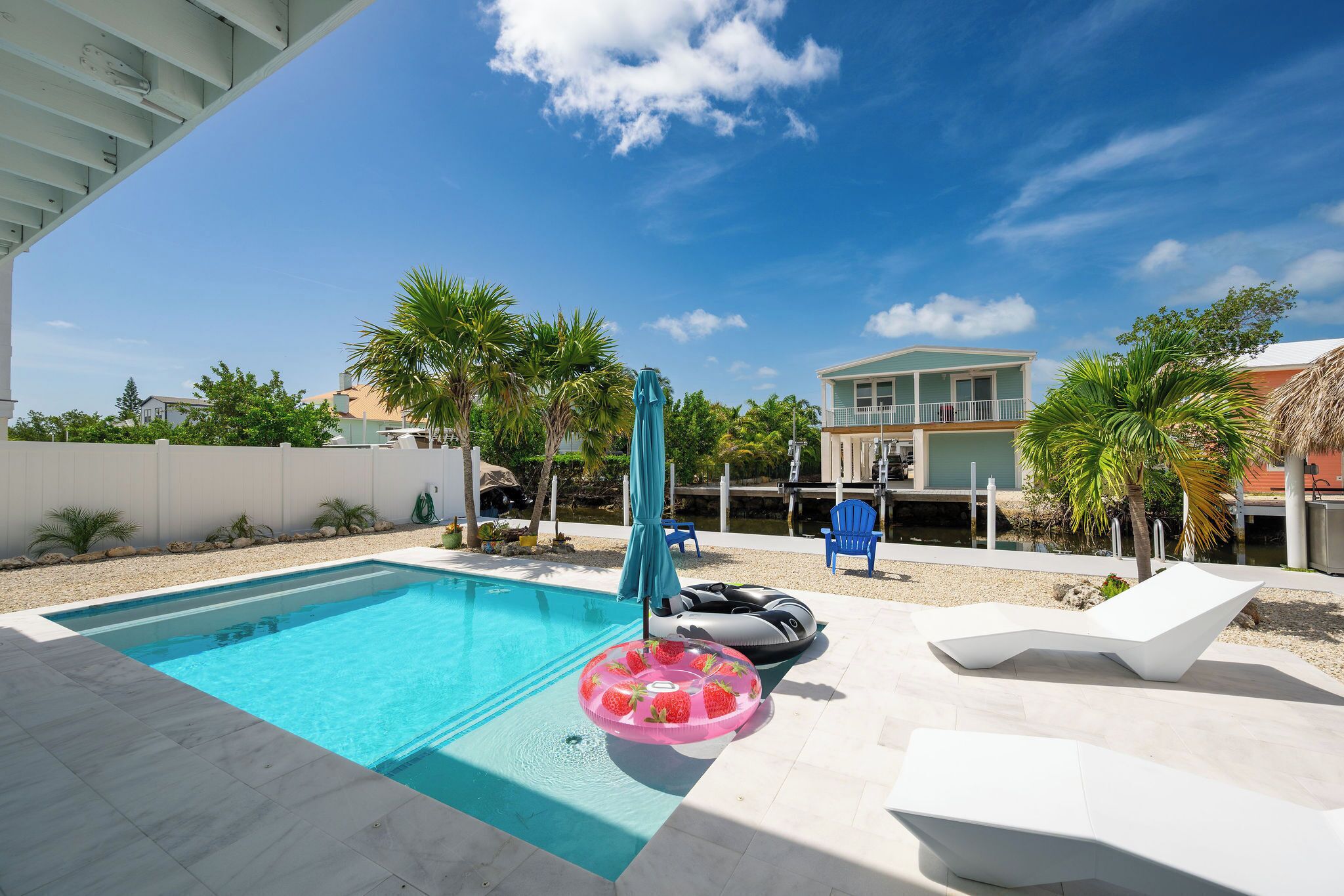A swimming pool with a clear blue water, surrounded by a white deck. There are lounge chairs and a blue umbrella.