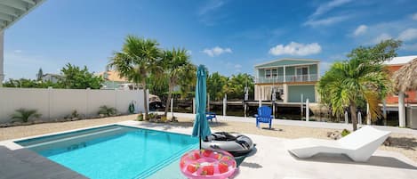 A swimming pool with a clear blue water, surrounded by a white deck. There are lounge chairs and a blue umbrella.