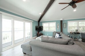 A spacious living room with a grey sofa, a ceiling fan, and large windows.