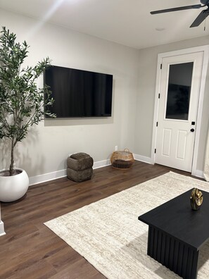 Ein Zimmer mit einem an der Wand befestigten Flachbildfernseher, einer Topfpflanze, einem runden Hocker, einem gewebten Korb, einem schwarzen Couchtisch und einer weißen Tür mit Glasfenster.