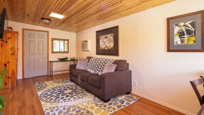 A living room with a brown sofa, a patterned rug, wooden flooring, a door, a small table, and a wall-mounted air conditioner. - General Sherman Loft ~ 2 min from Sequoia Nat’l Park (Three Rivers)