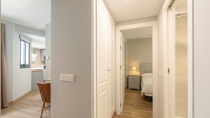Een hotelgang met een houten vloer, een eetgedeelte met een stoel en tafel, en een slaapkamer met een bed en nachtkastje.