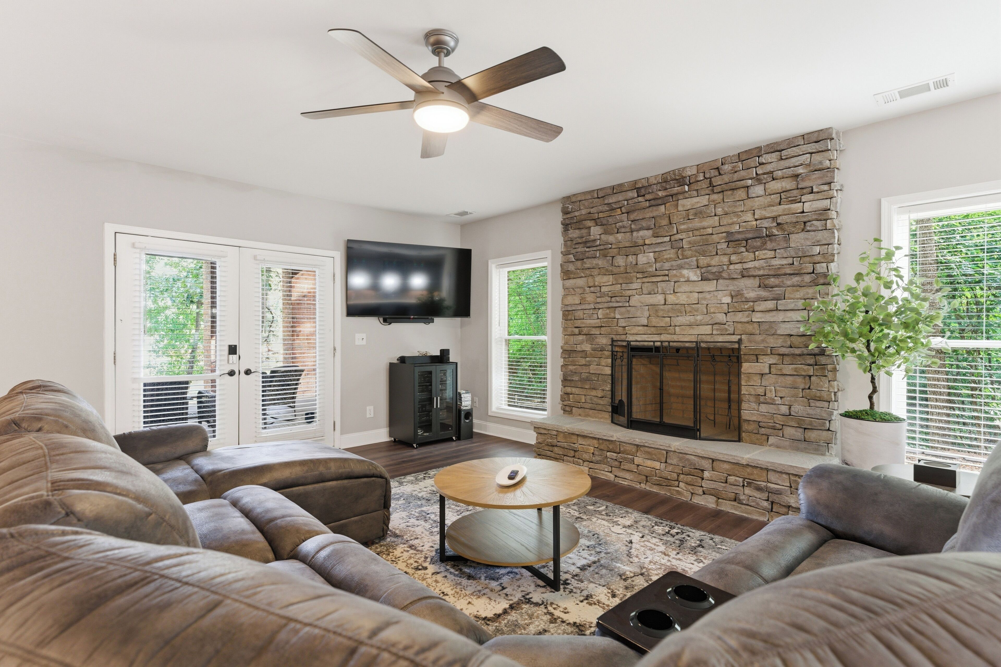 A living room with a stone fireplace, a ceiling fan, and a flat-screen TV mounted on the wall.