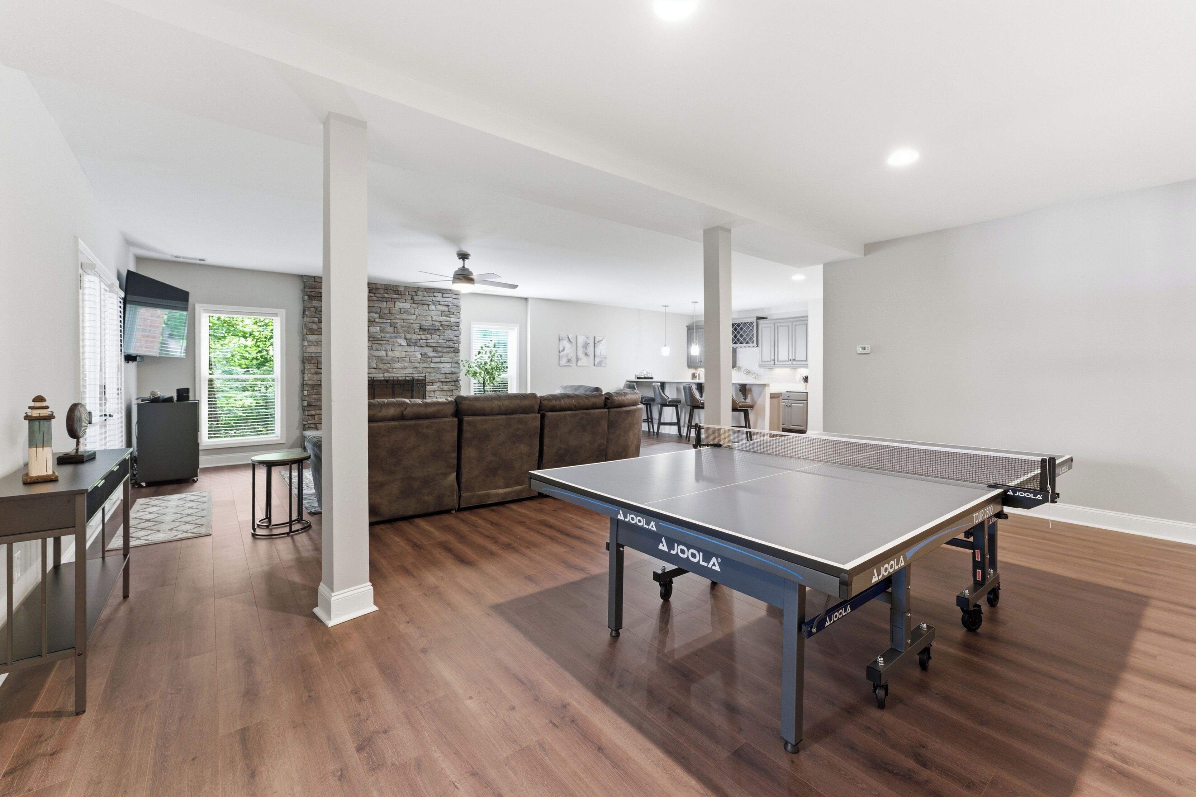 A spacious living room with a ping pong table, a stone fireplace, and a modern kitchen in the background.
