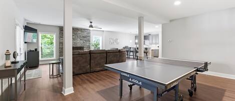 En rummelig stue med et pingpongbord, en pejs af sten og et moderne køkken i baggrunden.