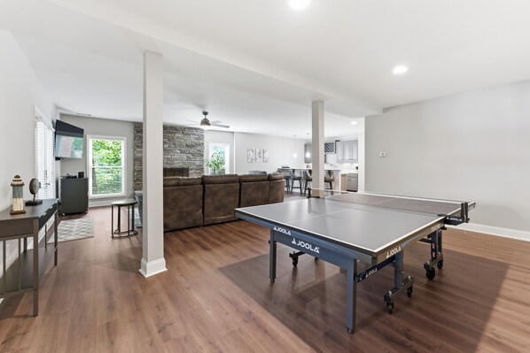 A spacious living room with a ping pong table, a stone fireplace, and a modern kitchen in the background.