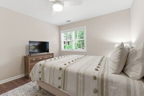 A bedroom with a bed, a television on a wooden dresser, a window with blinds, and a ceiling fan.