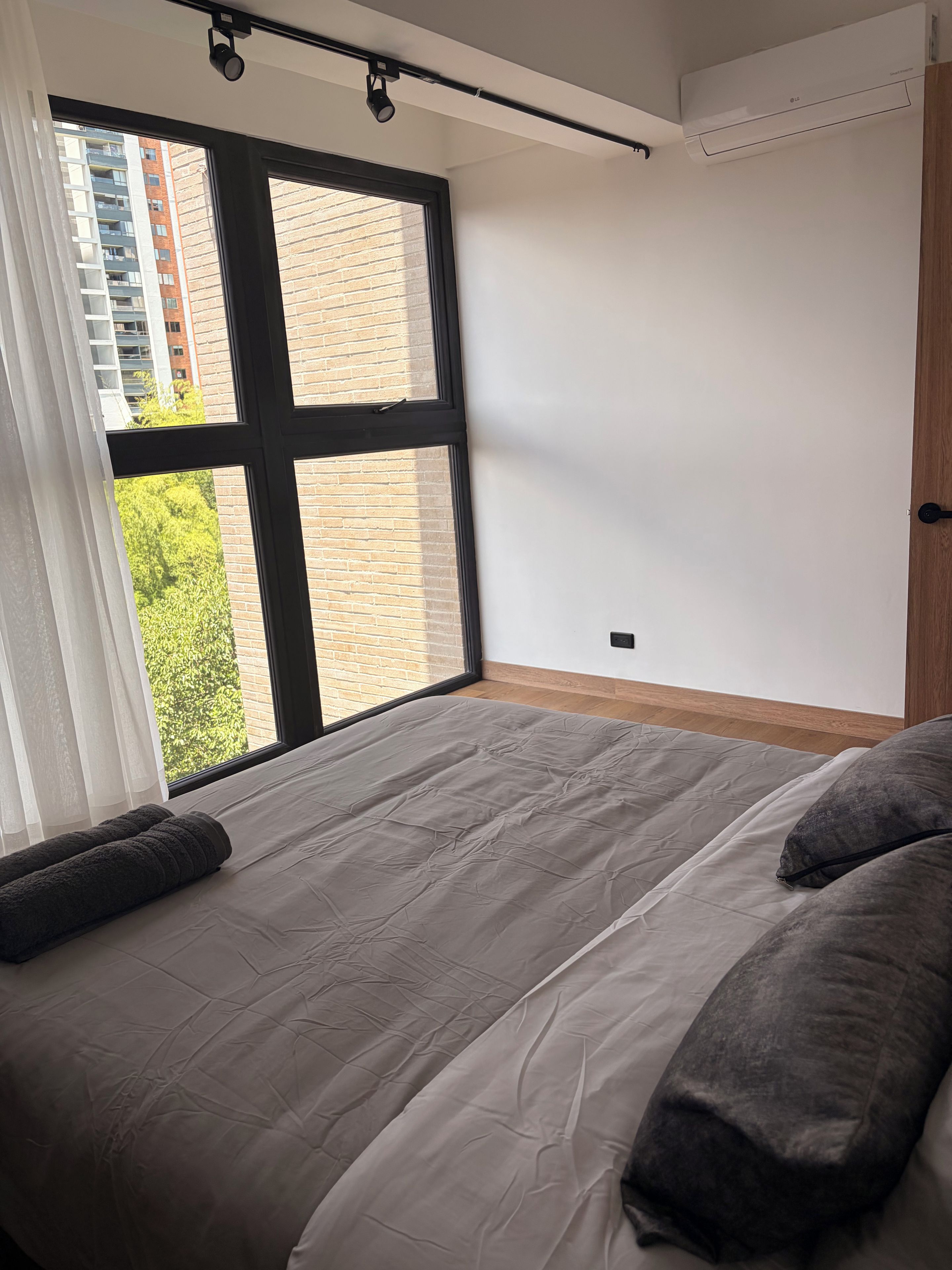 Un dormitorio con un ventanal grande, una cama con ropa de cama gris y piso de madera.