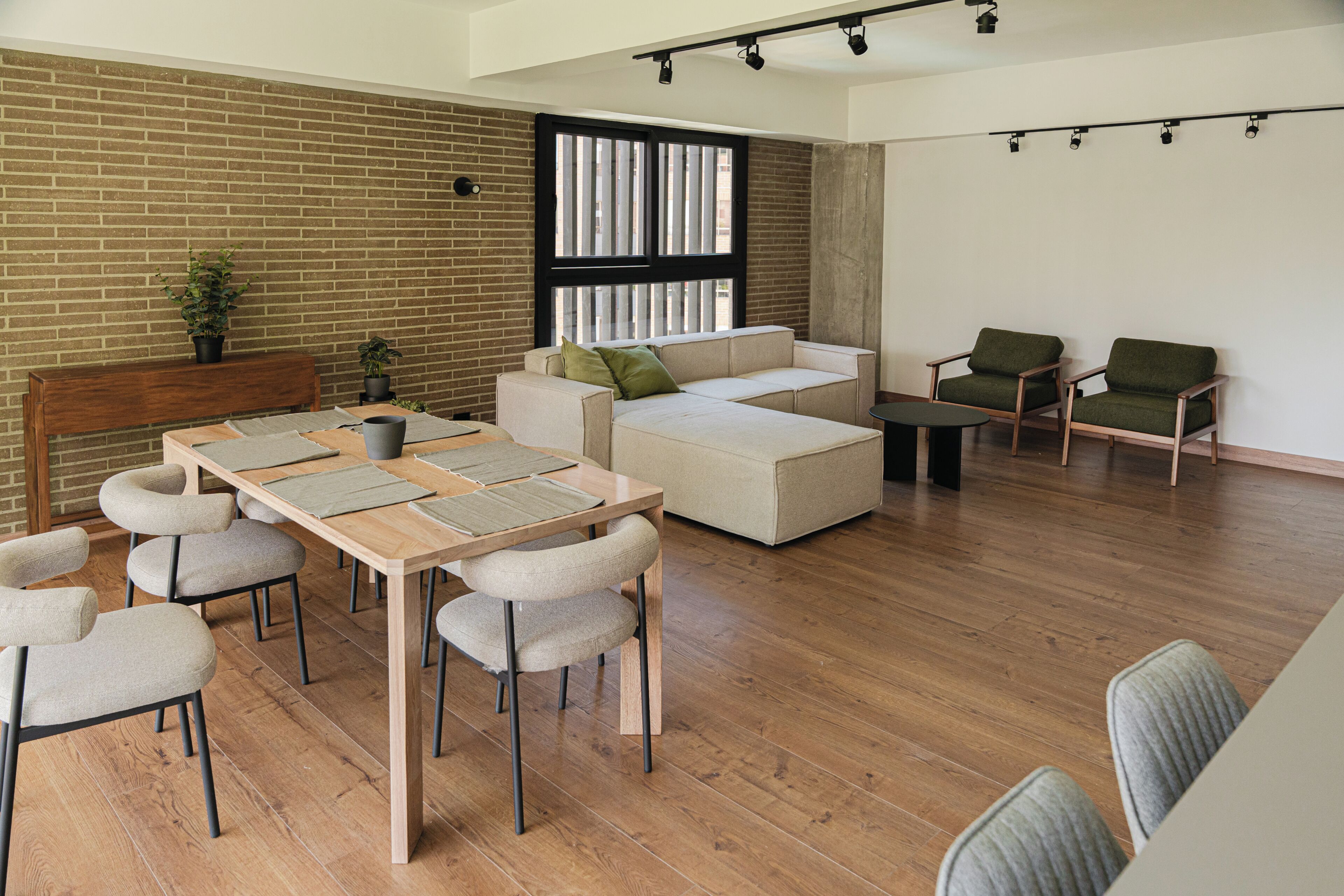 A spacious living area with a wooden floor, a sofa set, a dining table with chairs, and a brick wall.