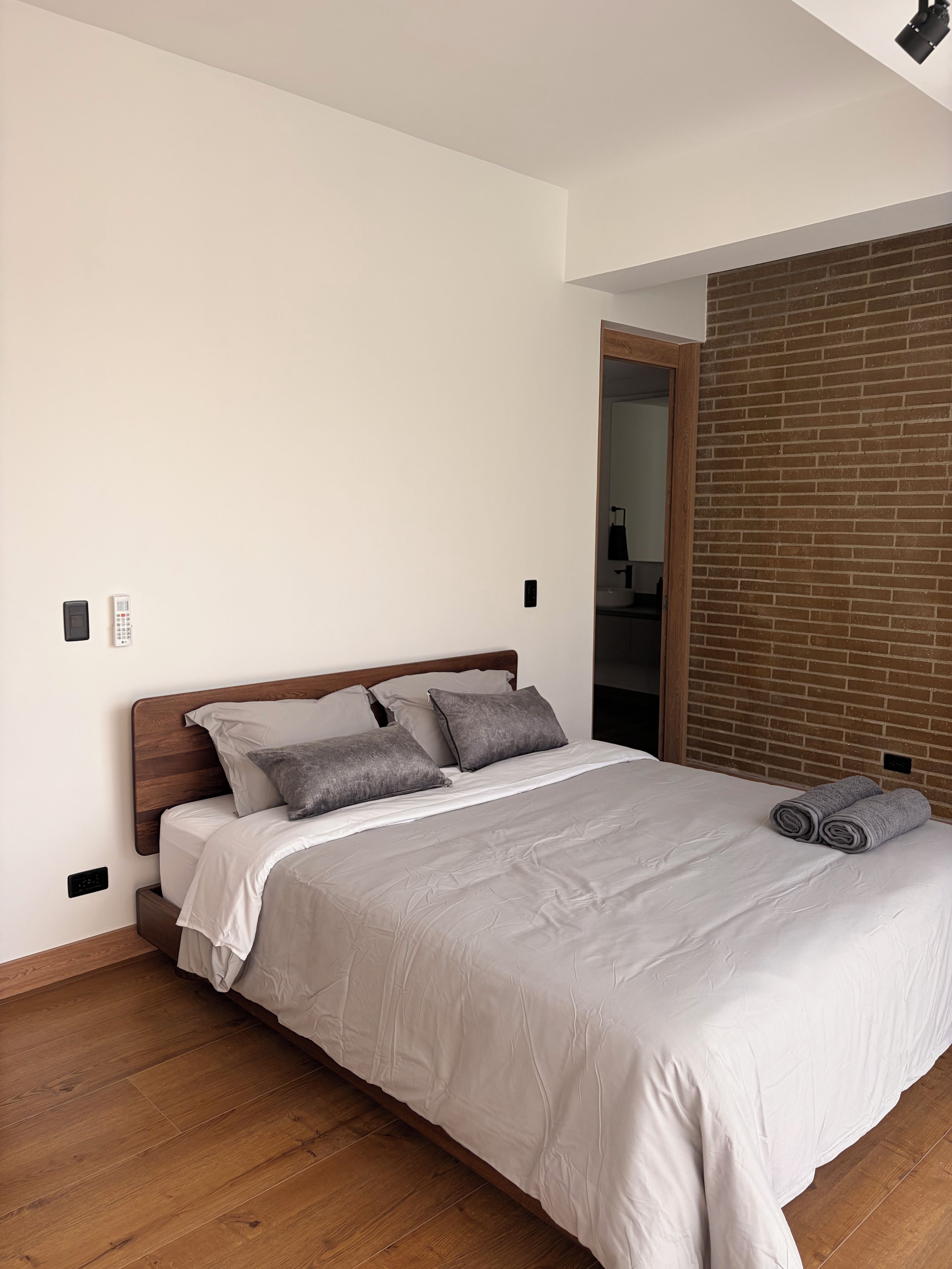 A neatly made bed with a wooden headboard, a brick wall, and a wooden floor.