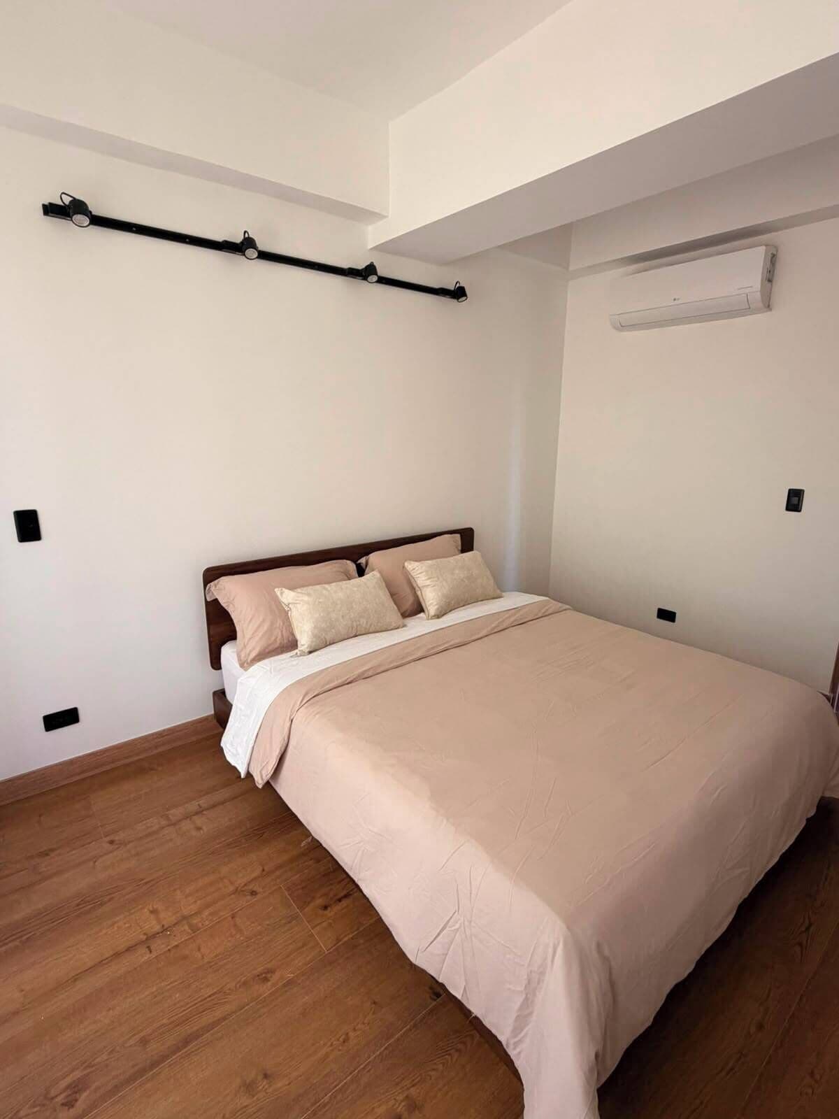 A bedroom with a bed, wooden flooring, a ceiling track lighting system, and an air conditioning unit.