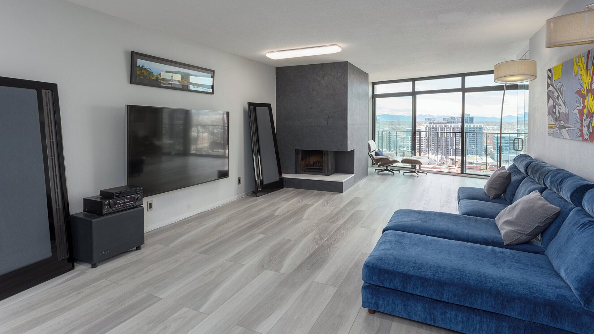 A modern living room with a flat-screen TV, a fireplace, a blue sofa, and a large window offering a city view.