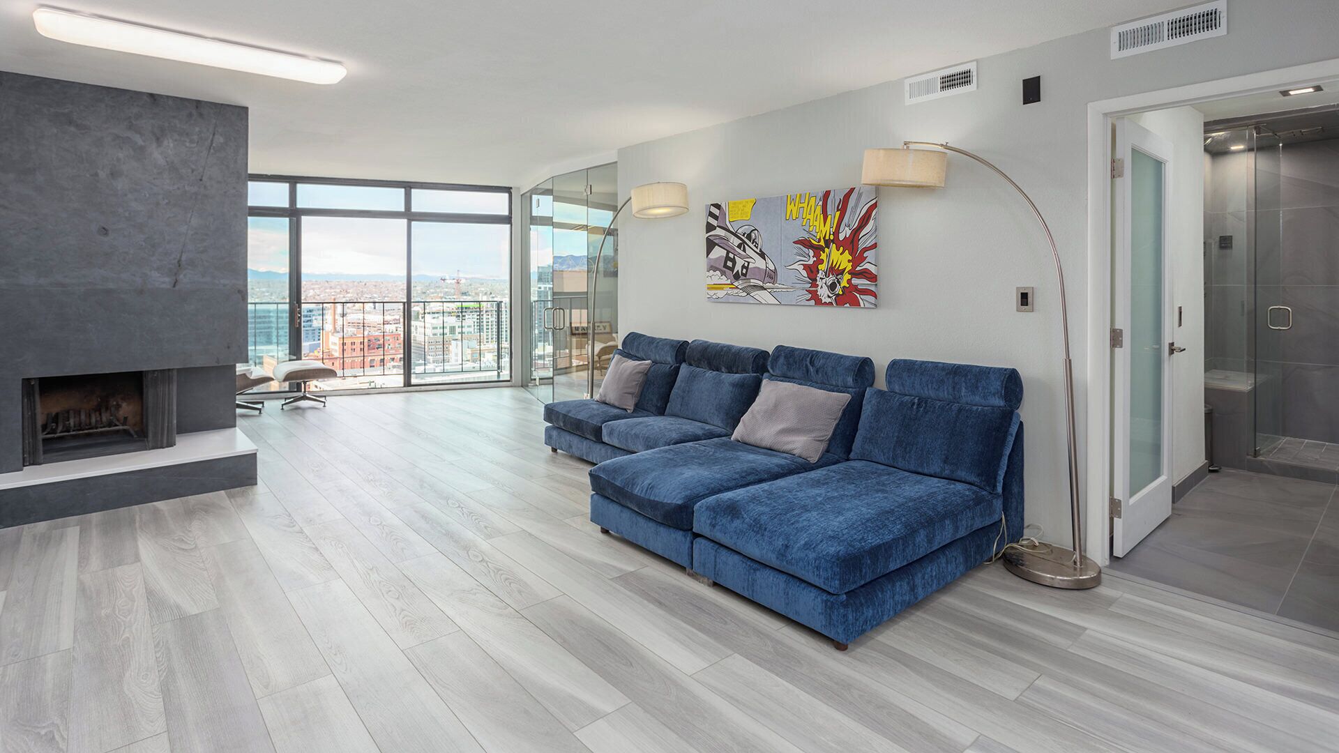A modern living room with a fireplace, a blue sofa set, a large window with a city view, and a glass shower enclosure.