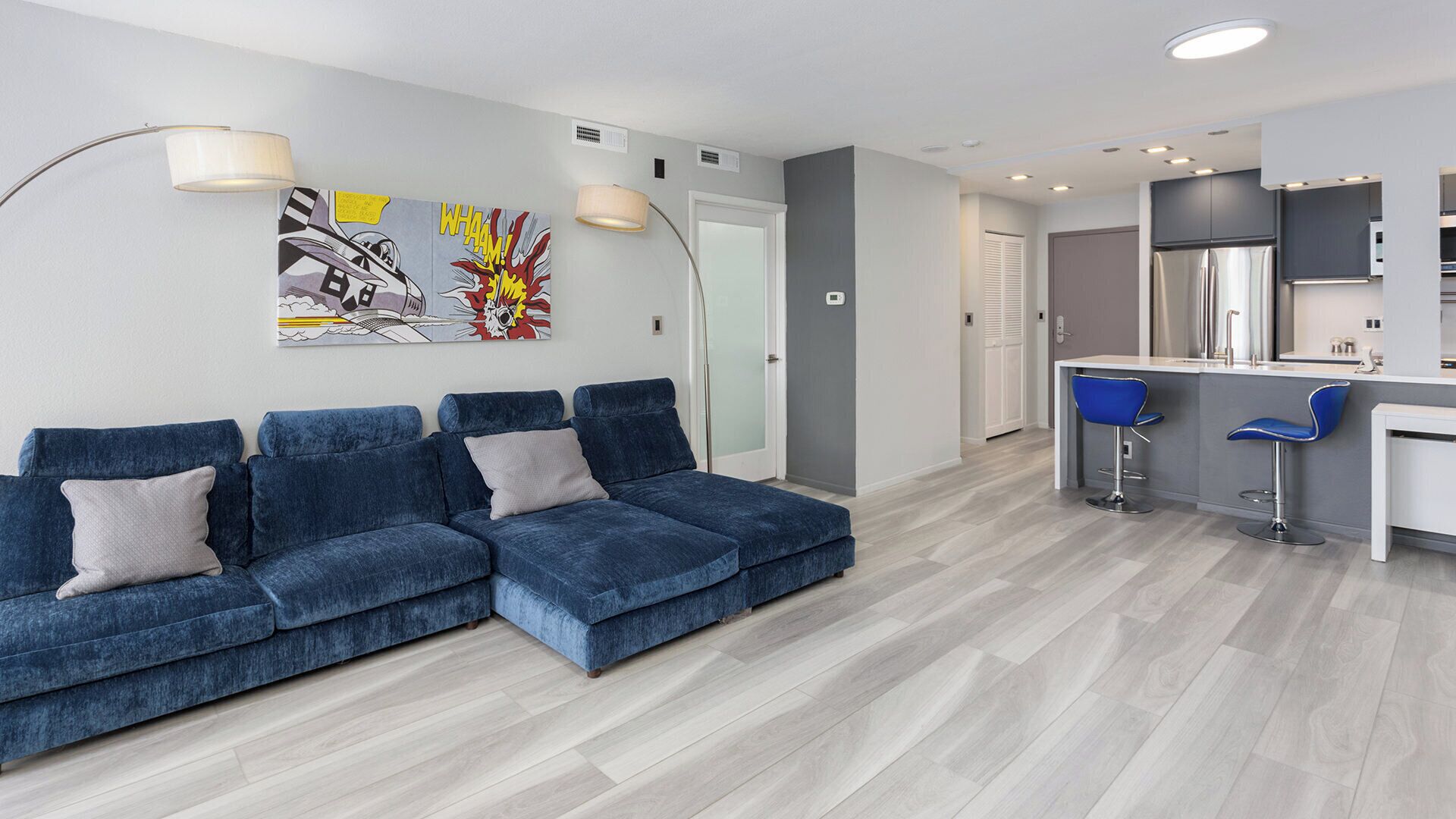 A modern living room with a sectional sofa, a dining area with bar stools, and a kitchen with stainless steel appliances.