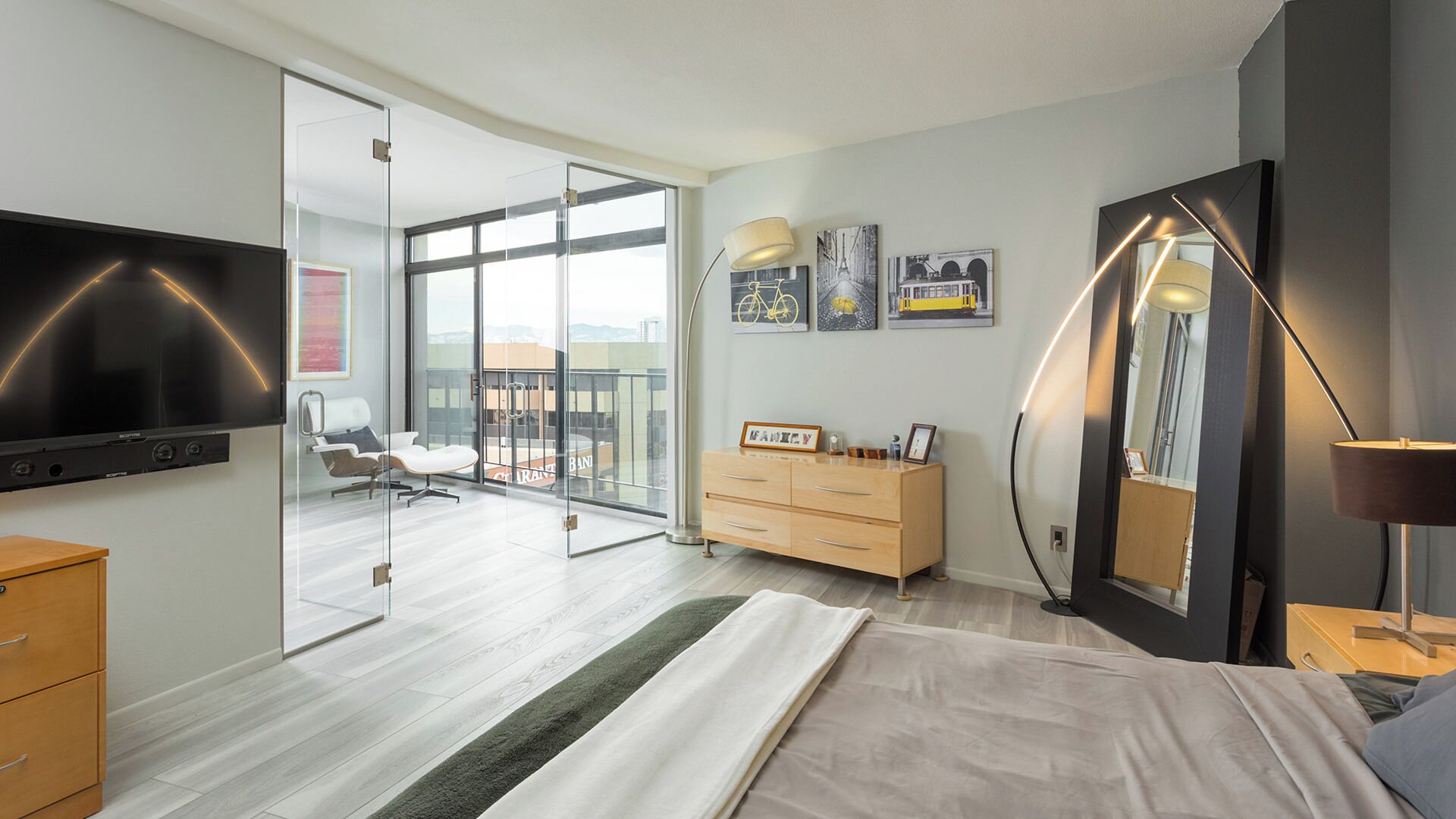 A modern bedroom with a large bed, a flat-screen TV, a dresser, and a balcony with a view.