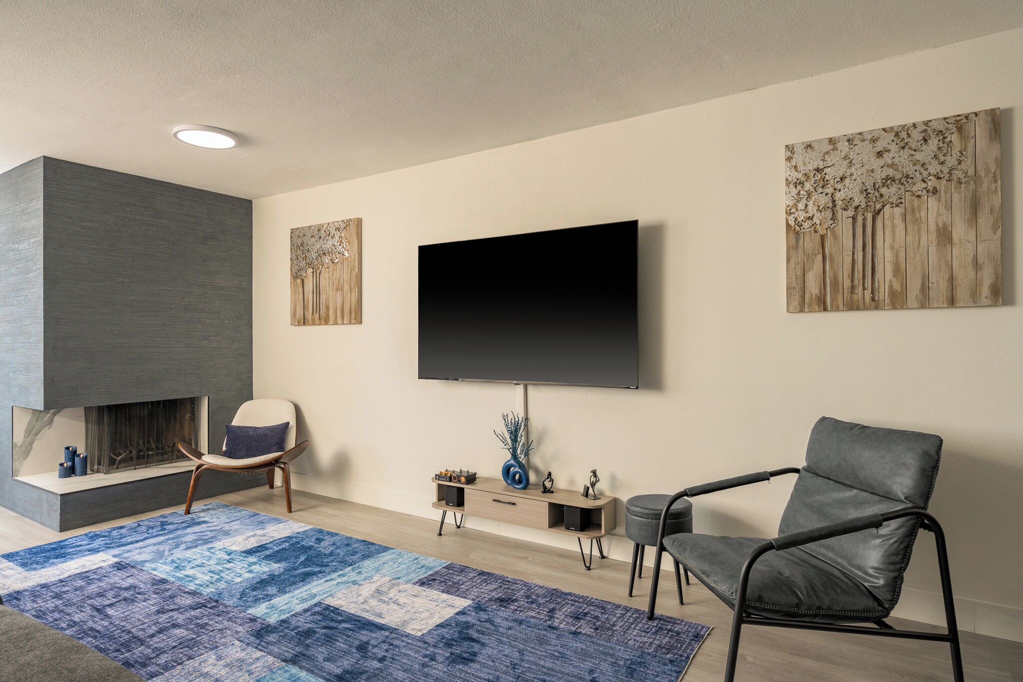 A modern living room with a flat-screen TV mounted on the wall, a fireplace, a blue patterned rug, and two chairs.
