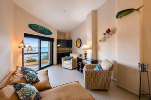 Ein Wohnzimmer mit einer Couch, Sesseln, einem Couchtisch und einem Fernseher. Eine Glastür führt auf einen Balkon mit Blick aufs Meer.
