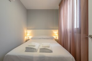 Ein Einzelbett mit weißen Bettwäsche und zwei gefalteten Handtüchern, zwei Nachttischlampen und eine Tür mit Vorhang.