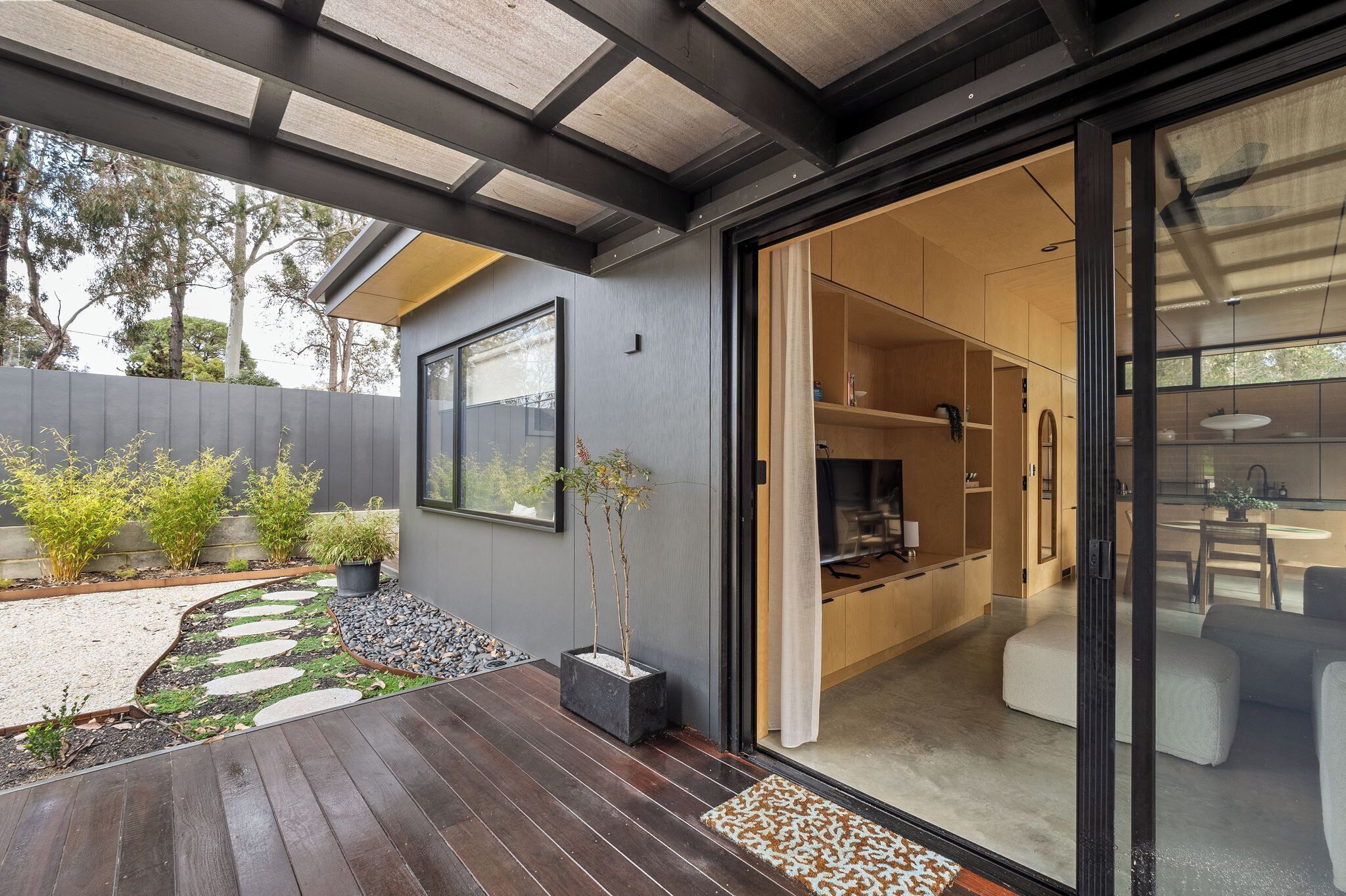 Een modern huis met een houten terras, een tuin met staptegels en een schuifdeur die leidt naar een keuken.