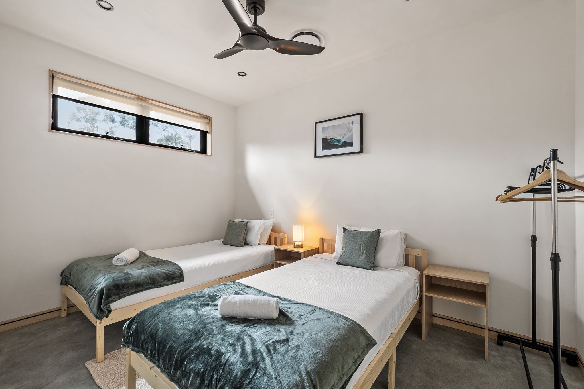 A bedroom with two beds, a ceiling fan, a window, a bedside table, a lamp, and a coat rack.