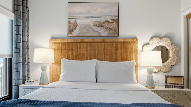 Habitación de hotel con una cama grande, dos lámparas de noche, un espejo decorativo y un cuadro de una playa.