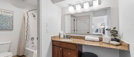A bathroom with a toilet, shower, vanity with a mirror, and a trash bin.