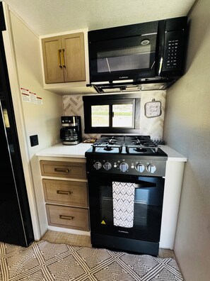 A compact kitchenette with a stove, microwave, coffee maker, and a small window. - 2-bedroom recreational vehicle in lovely Exeter with AC (Exeter)