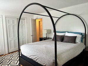 Ein Himmelbett mit einem gemusterten Überwurf, ein Nachttisch mit Lampe und ein Teppich mit geometrischem Muster.