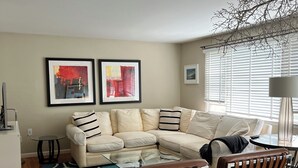A modern living room with a sectional sofa, a glass coffee table, a patterned rug, and a white shelving unit.