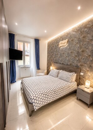 Un dormitorio moderno con una cama grande, un televisor de pantalla plana, una mesita de noche con lámpara y una ventana con cortinas.