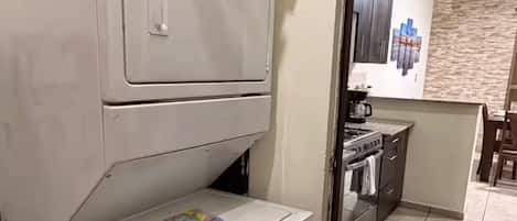 A compact laundry room with a washing machine and dryer, a small table with a book, and a kitchen area visible through an open door.