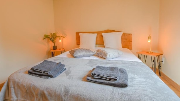 Uma cama bem arrumada com almofadas e toalhas dobradas, uma planta na mesinha de cabeceira e um criado-mudo com abajur.